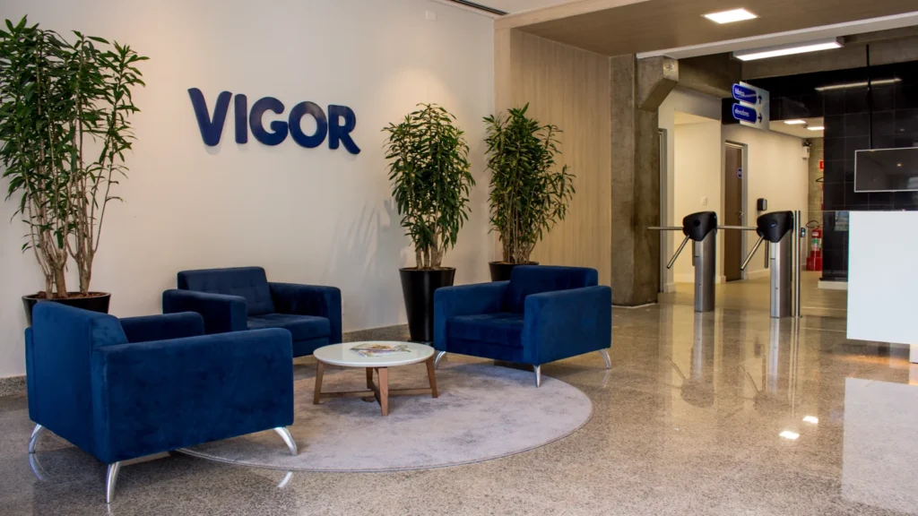 Recepção da fábrica com o logotipo Vigor na parede, poltronas azuis e catracas ao fundo